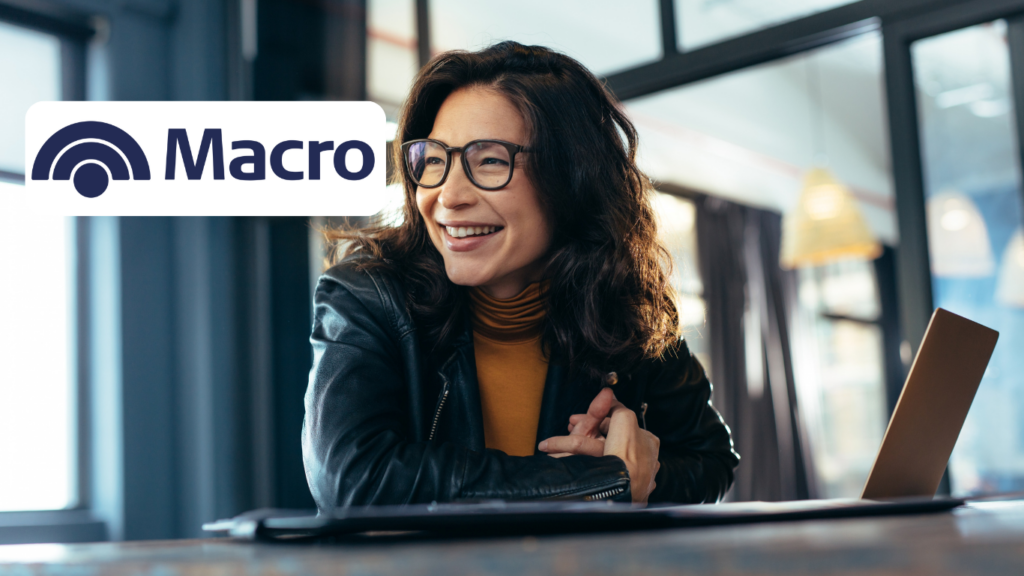 Una mujer sonriente, con gafas y chaqueta de cuero, sentada frente a una computadora portátil en un ambiente de oficina. Ella parece estar disfrutando de una conversación o de una interacción en línea, y está vestida con una camiseta de color mostaza. En la esquina superior izquierda, aparece el logo de Macro.