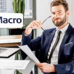 Un hombre con barba y gafas, vestido con un traje formal, sonríe mientras sostiene un documento en una mano y unos lentes en la otra. Está sentado en una mesa de oficina con una laptop y documentos alrededor. En la esquina superior izquierda, aparece el logo de Macro.